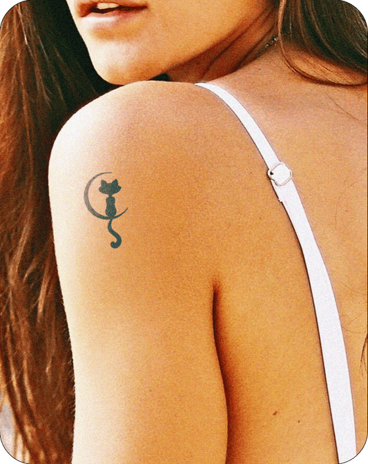 A woman with long brown hair and a white spaghetti strap top shows a minimalist black tattoo of a cat sitting inside a crescent moon, placed on her upper shoulder.