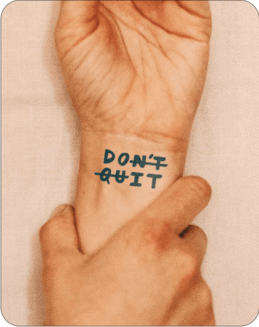 Tattoo on the wrist with the phrase 'DON’T QUIT,' with the word 'QUIT' crossed out, accompanied by a hand gently holding the wrist.