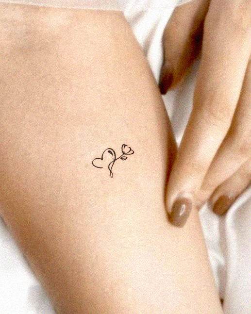 A simple, elegant tattoo of a heart and flower on a person's forearm, with a hand gently resting beside it. The tattoo features a thin, black outline of a heart and a small flower stem. The person's nails are painted a neutral shade.
