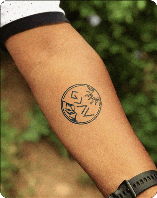 Alt text: A tattoo on the inner forearm showcasing a circular design. The tattoo features a stylized sun with rays at the top and a mountain range at the bottom. The word "GEAV" is written in bold, angular letters within the circle, positioned between the sun and mountains. The tattoo is inked in dark blue, contrasting against the skin, and the background shows blurred greenery.
