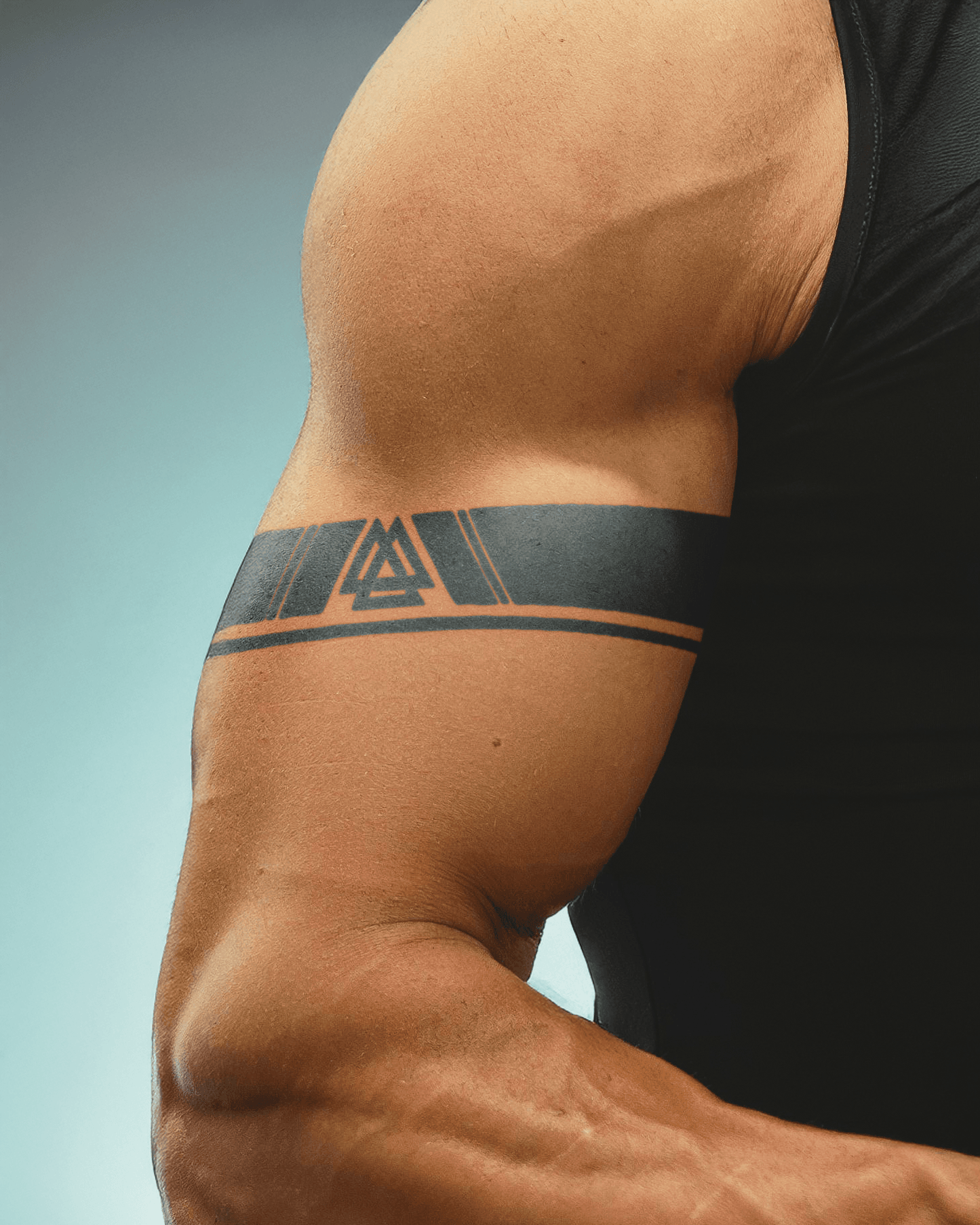 A muscular upper arm displaying a black armband tattoo featuring a bold geometric design with a central Valknut symbol—three interlocking triangles—flanked by sharp angled accents and bordered by two parallel curved lines.