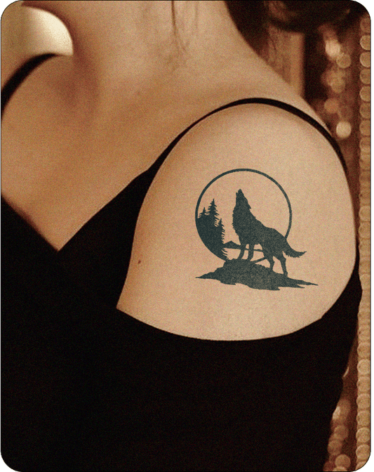 A dark silhouette tattoo of a howling wolf set against a circular backdrop, possibly representing the moon or sun, is applied to the upper arm of a person. The wolf stands on a textured ground, and stylized pine trees are visible within the circle, creating a scene of wilderness. This design suggests a semi-permanent tattoo style suitable for Inkhub, emphasizing a bold, graphic aesthetic.