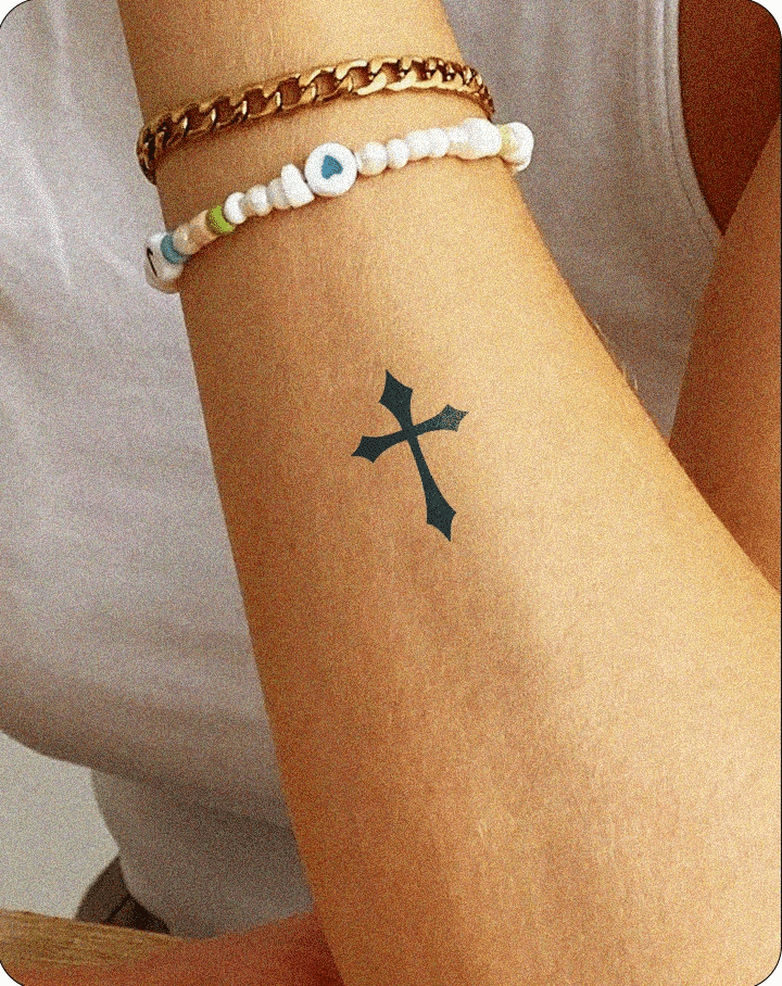  A semi-permanent tattoo of a black Gothic-style cross on the inner forearm of a person, shown wearing a white tank top and two bracelets—one gold chain and one beaded with a heart charm—resting casually on a wooden surface.