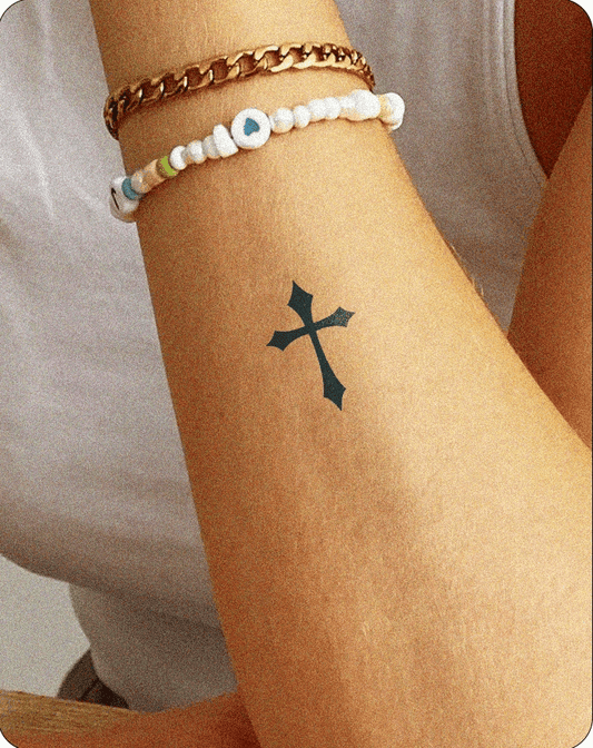  A semi-permanent tattoo of a black Gothic-style cross on the inner forearm of a person, shown wearing a white tank top and two bracelets—one gold chain and one beaded with a heart charm—resting casually on a wooden surface.