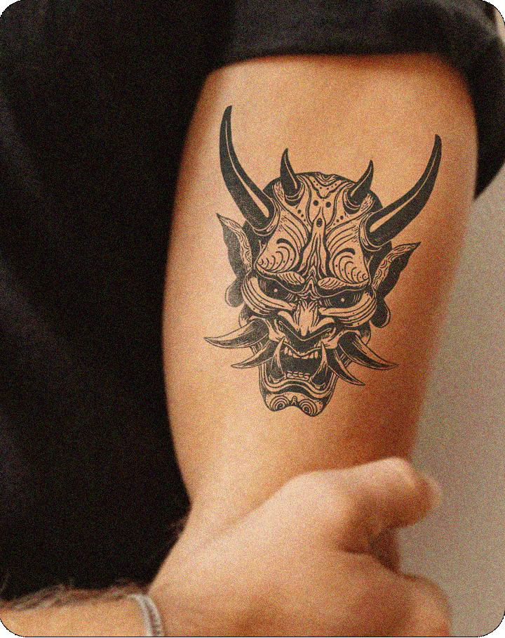 ChatGPT said:
The image shows a detailed tattoo of a demonic mask on the upper arm of a person. The tattoo features a traditional Japanese Oni mask, with large, sharp horns and fierce, snarling features. The mask’s intricate designs include swirling patterns on the forehead and chin, enhancing its fierce and ornate appearance. The intense eyes, elongated tusks, and sharp lines give the tattoo a strong, aggressive look. The arm is partially visible, with the person holding their wrist, adding a dynamic pose 