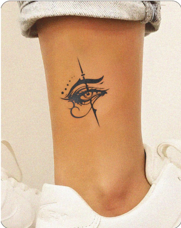 A close-up of an ankle with light tan skin features a striking semi-permanent tattoo of a stylized eye. The design incorporates fine lines, bold accents, and dot work, blending ancient Egyptian influences with a modern twist. The wearer sports white sneakers and rolled-up light denim, reflecting InkHub’s blend of artistic edge and everyday wearability.