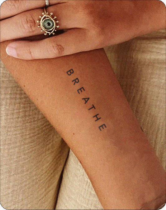 A close-up of a forearm with medium-tan skin features a semi-permanent tattoo of the word “BREATHE” in clean, black capital letters arranged vertically. A hand rests gently across the arm, wearing a detailed ring with an eye-like design. The tattoo’s minimal and calming style reflects InkHub’s focus on intentional, everyday wearable art.