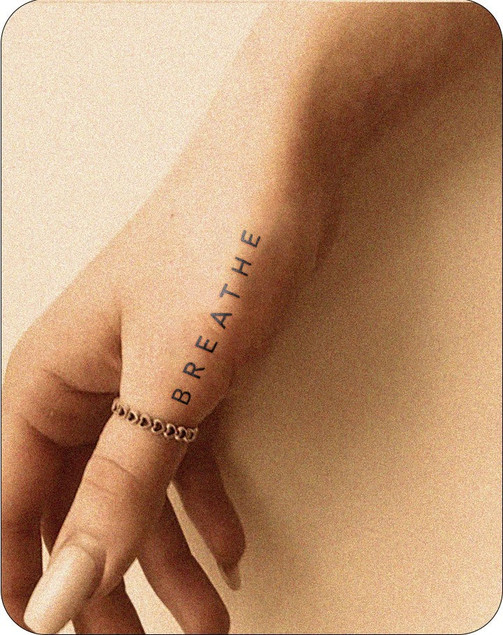 A close-up of a hand with a neutral manicure and silver chain ring shows a semi-permanent tattoo along the outer side of the hand. The black ink reads “BREATHE” in clean, all-caps vertical lettering. The warm-toned lighting and soft shadows give the image a calming, mindful atmosphere, echoing InkHub’s blend of simplicity and self-expression.