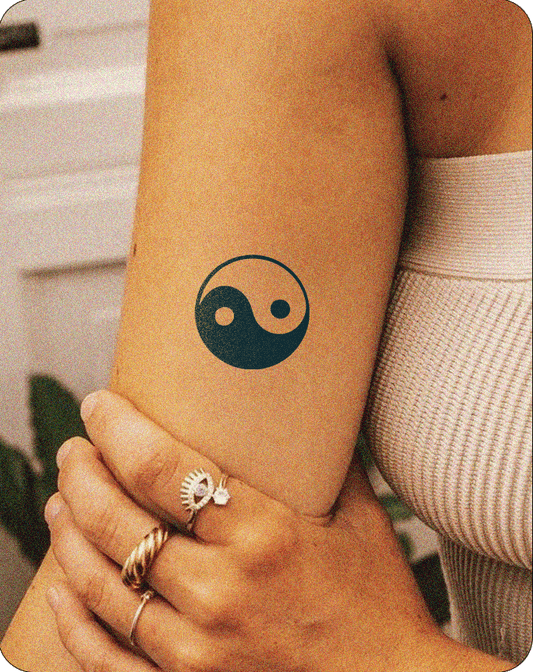 Yin Yang symbol tattooed on the inner forearm of a person wearing a sleeveless knit top and gold rings.