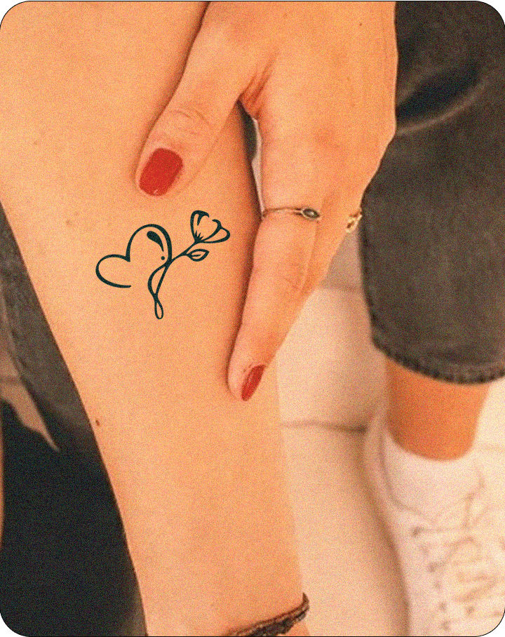 A minimalistic tattoo of a heart and flower on a person's forearm, with a hand gently touching it. The tattoo features a fluid design with a simple flower stem. The person wears red nail polish and a silver ring.

