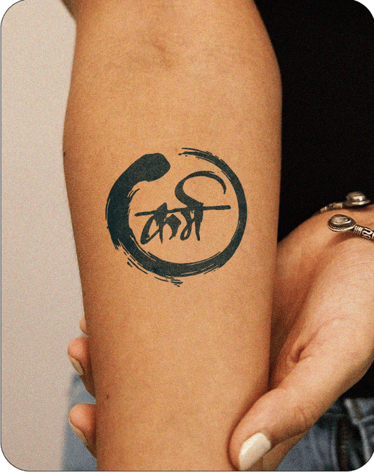 A black semi-permanent tattoo design featuring the Hindi word "कर्म" (karma) written in a bold, expressive brush script. The calligraphy is enclosed within an enso-style circular brushstroke, symbolizing mindfulness and the cyclical nature of life. The overall design blends Eastern philosophy with modern minimalism