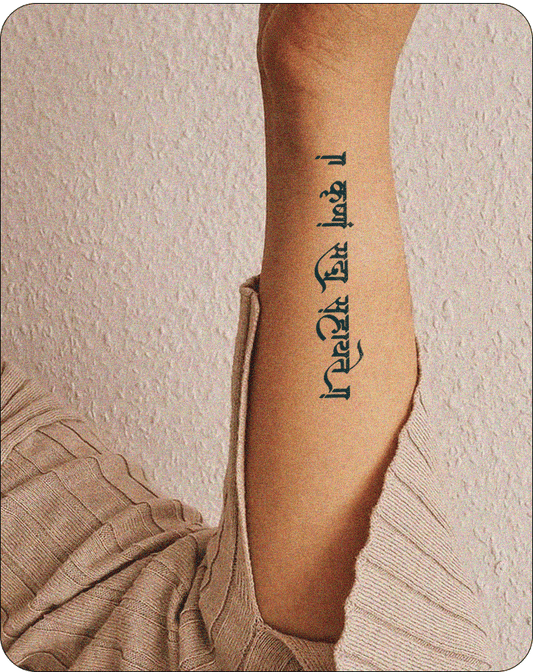  A forearm tattoo in Sanskrit script, written vertically in black ink, displaying the phrase "॥ कृष्णं सदैव सहायते ॥" on a person's inner arm, with the arm slightly bent and dressed in a cozy beige knit sleeve against a textured wall background.