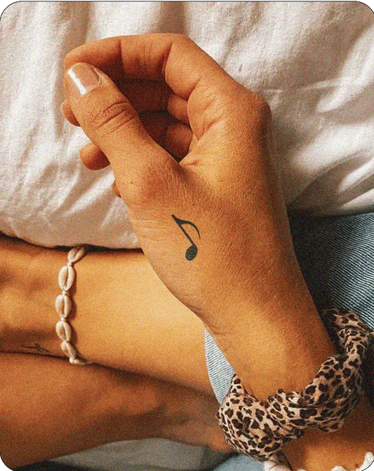 A small, black, semi-permanent tattoo from Inkhub, depicting an eighth note (quaver) musical symbol, is positioned on the back of a person's hand. The tattoo is located near the base of the thumb and is clearly visible against the person's skin. The person is also wearing a bracelet made of small, white, shell-like beads and a patterned scrunchie around their wrist.