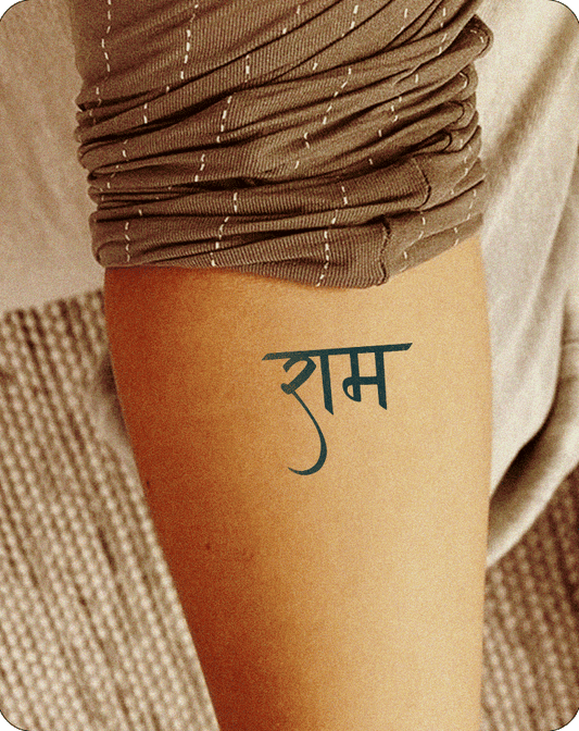 A dark teal, semi-permanent tattoo, likely from Inkhub, depicting the Hindi word "राम" (Ram) written in a stylized Devanagari script, is positioned on a person's inner forearm. The tattoo is clearly visible against the person's skin, which is partially covered by a brown, striped fabric.